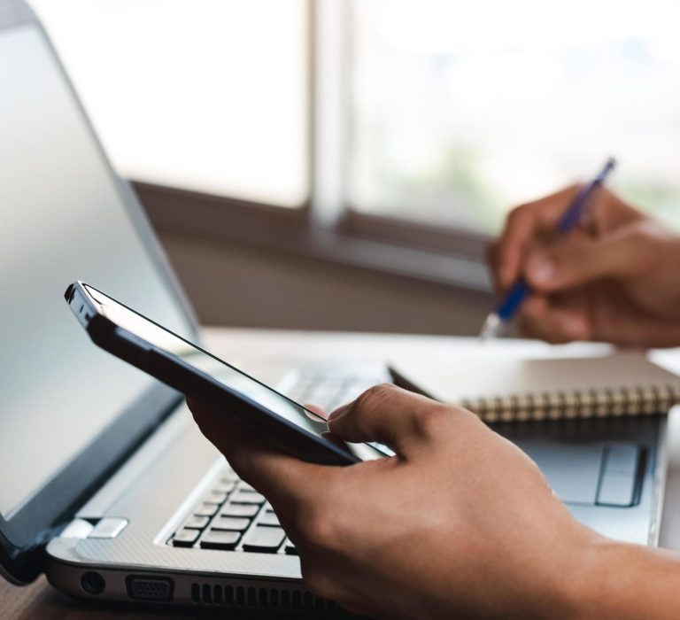 Young-Man-Using-Laptop-Computer-And-Mobile-Phone-When-Looking-For-Financial-Information-In-Business-Work-At-The-Desk-Writing-With-A-Pen-Studying-Remotely-From-Home-And-Working-From-Home-Free-Photo
